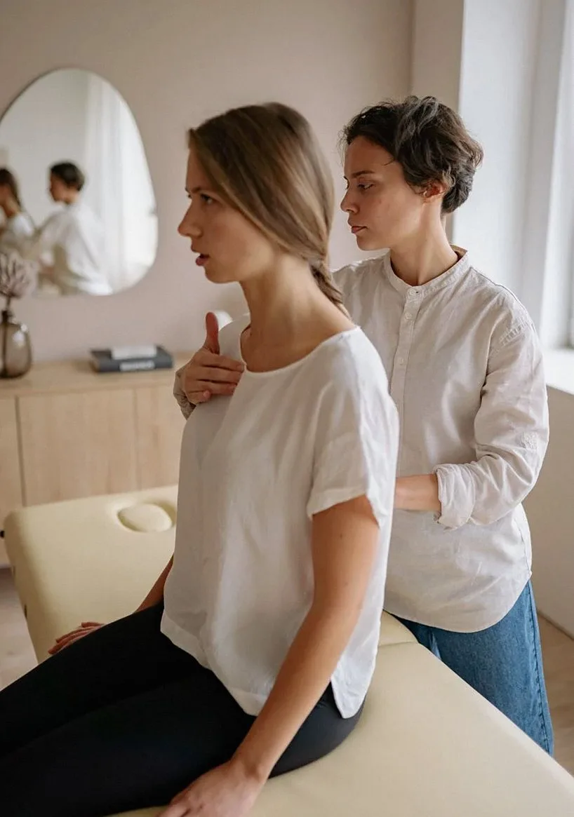 Two women in a bright room, one sitting on a treatment table while the other works on her shoulders