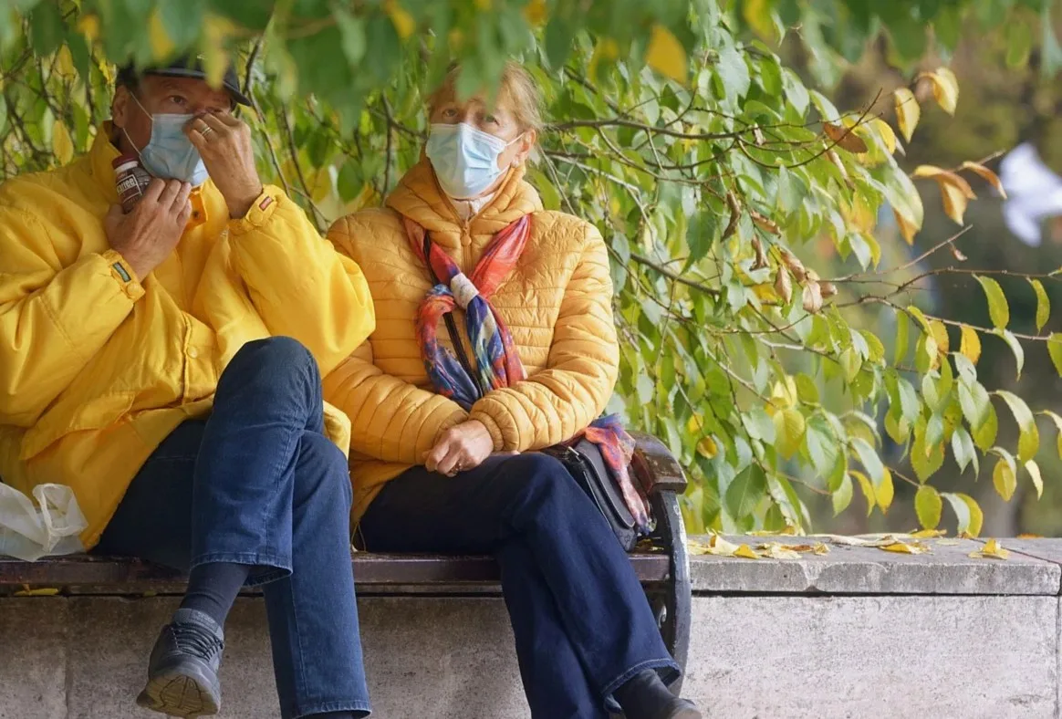 Two people in yellow jackets and face masks sitting on a bench under trees